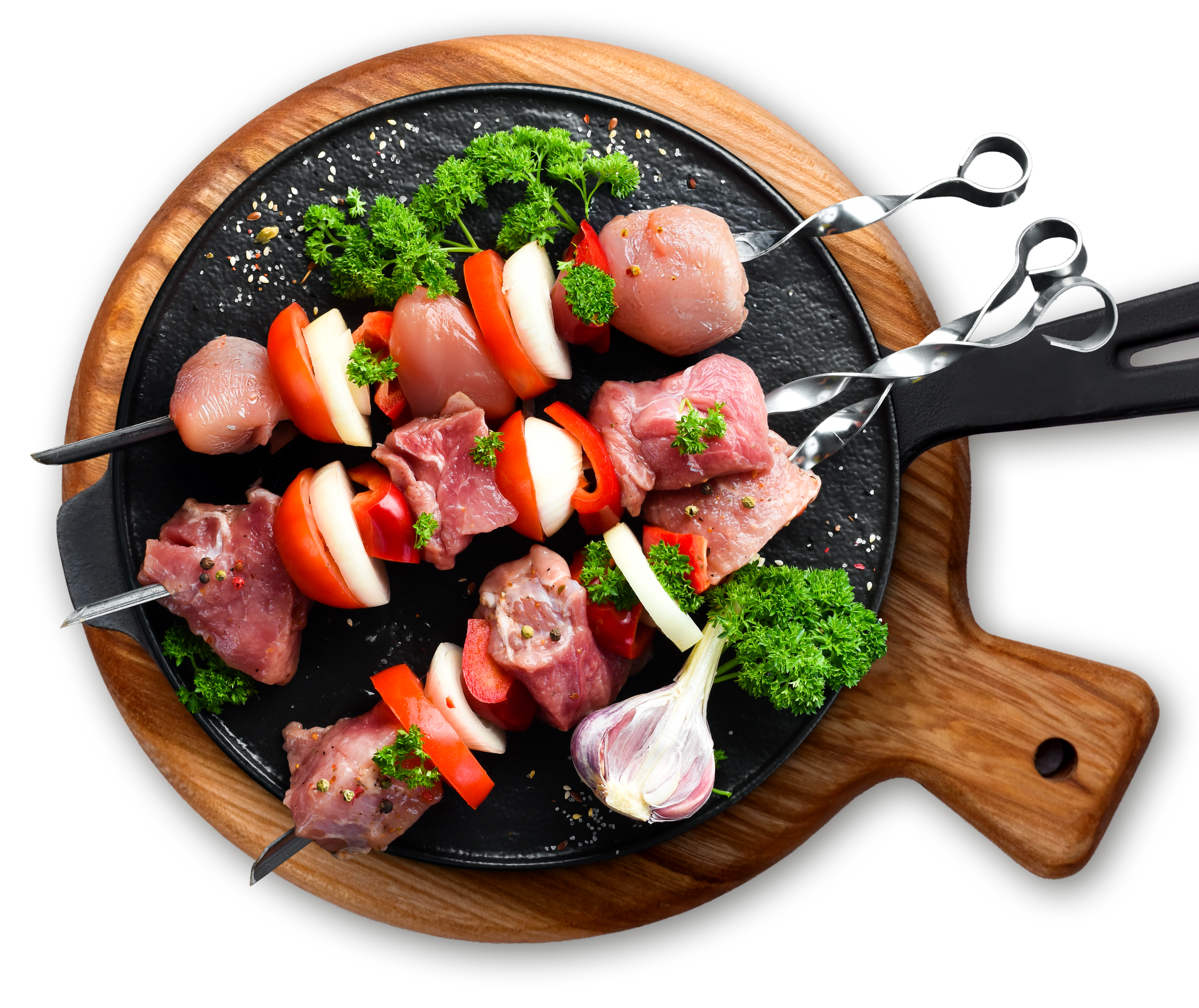 Surowe szaszłyki z kawałkami mięsa, papryki, cebuli i natki pietruszki, ułożone na czarnej patelni z drewnianą rączką, na desce do krojenia. W tle widoczna główka czosnku i przyprawy.